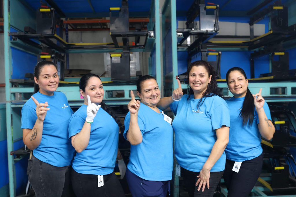 Plasman employees holding up number 1 finger in front of storage bins in Greer